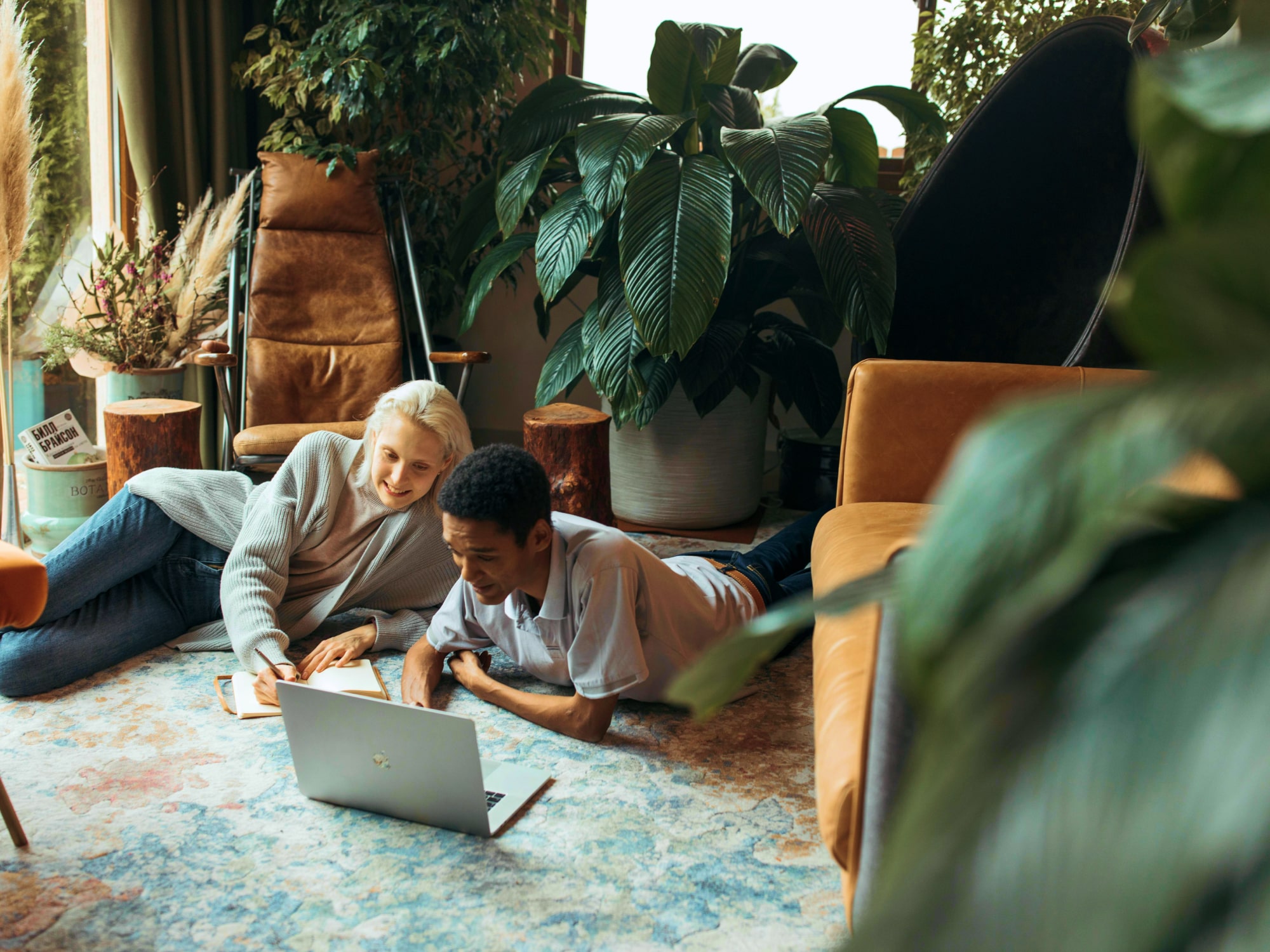 Two people lying on a rug with a laptop, surrounded by plants and cozy furniture.