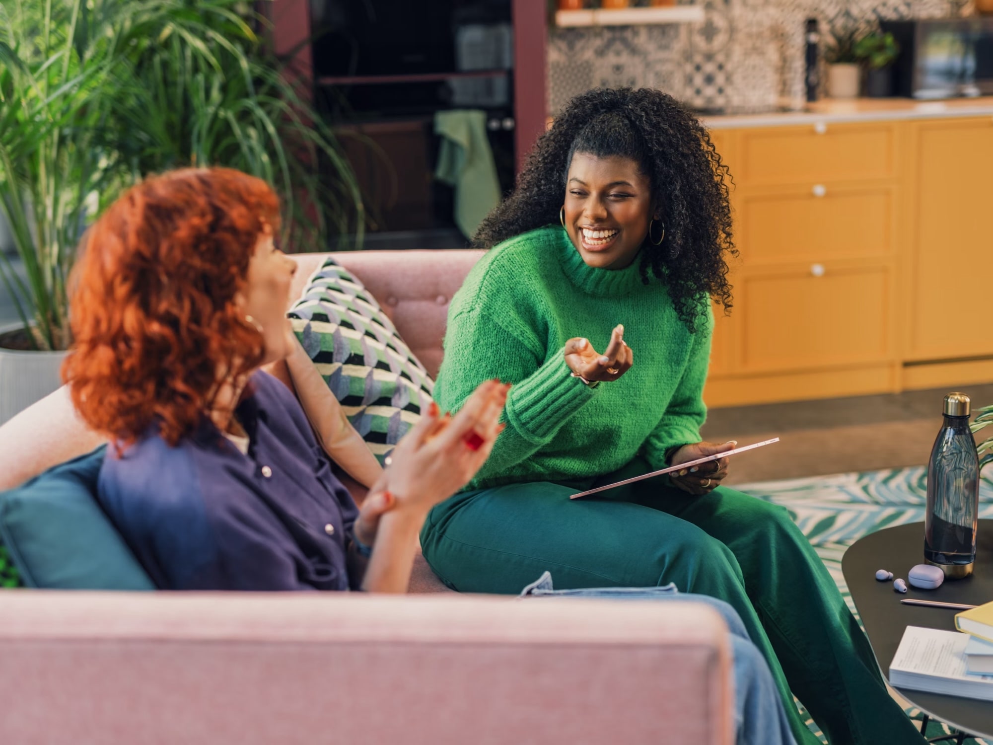 Two women smiling and talking on a pink sofa in a cozy, colorful living room setting.
