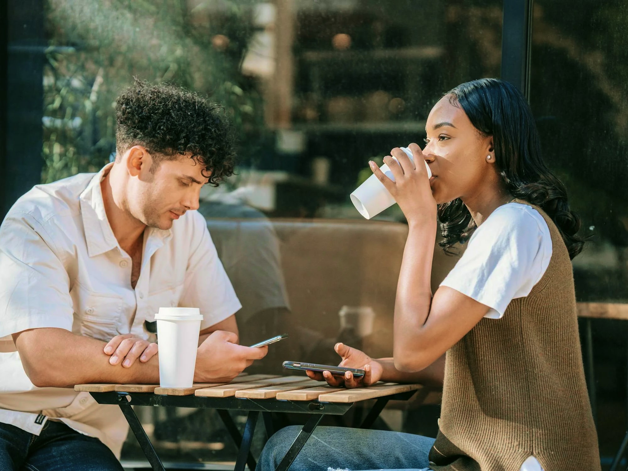 Man and woman sitting at a wooden table outside cafe, man looking at phone, woman drinking from cup.