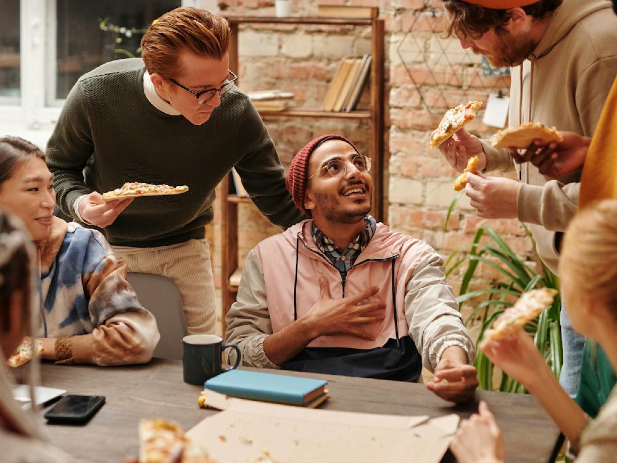 Group of diverse young adults eating pizza and chatting happily in a casual indoor setting.