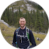 Smiling man wearing a backpack outdoors with trees and rocky hills in the background.