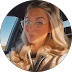 Woman with long blonde hair wearing clear-framed glasses and a beige jacket in a car.