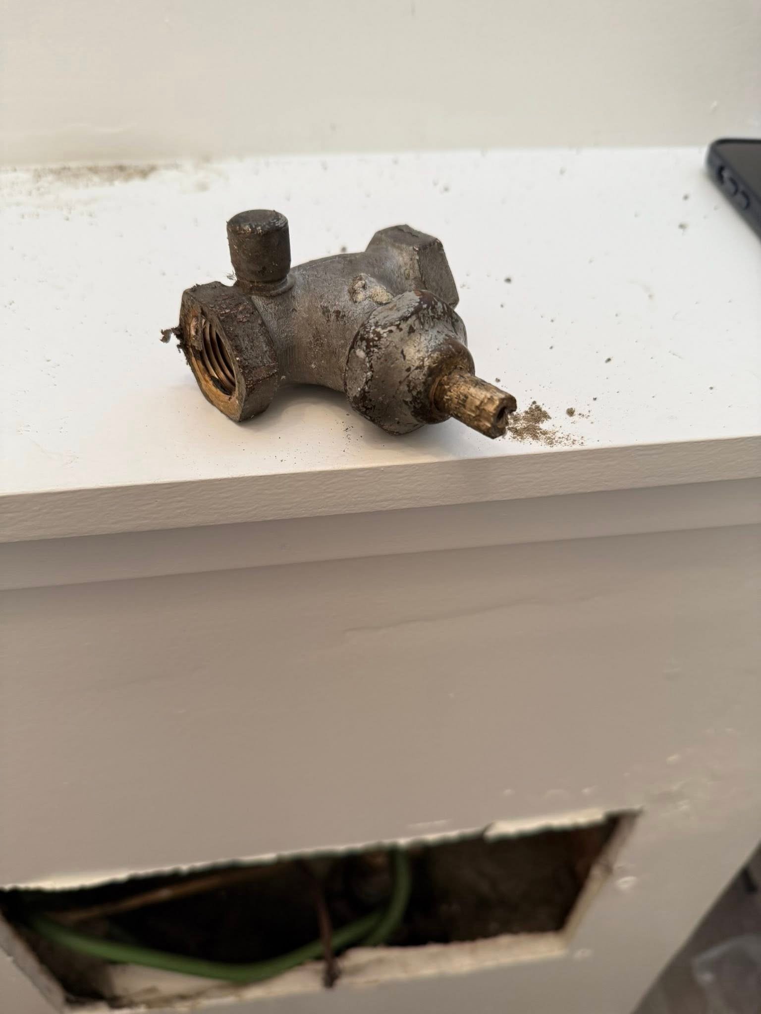 Old, rusty metal valve on a white surface with a rectangular hole below showing pipes inside.