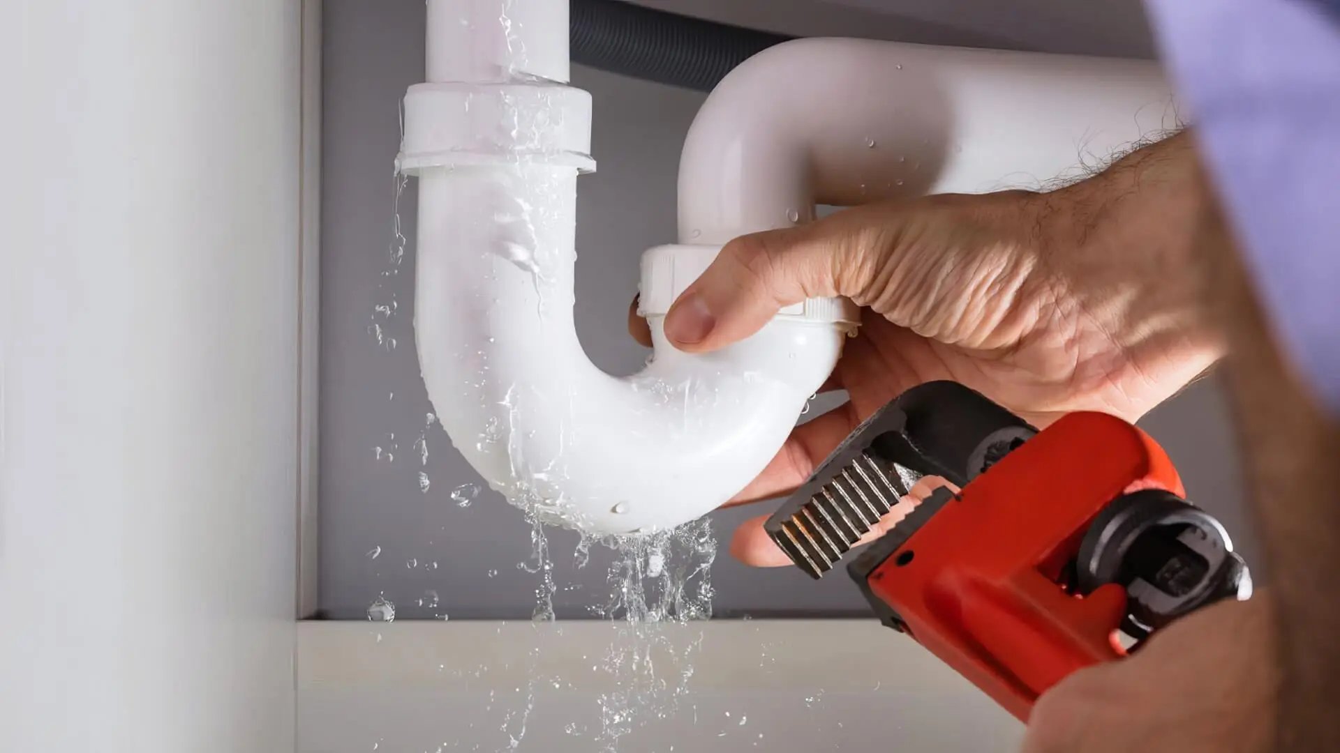 Hand holding a pipe wrench fixing a leaking white pipe under a sink with water dripping.