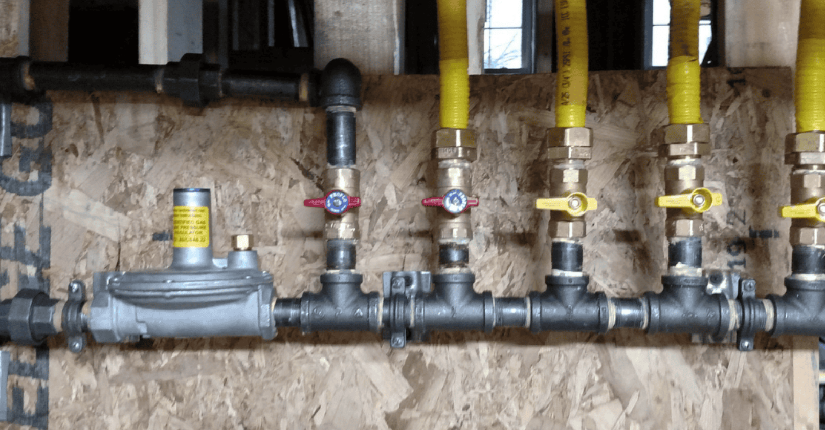 Black metal gas pipes with yellow valves and hoses mounted on wood paneling in utility area.