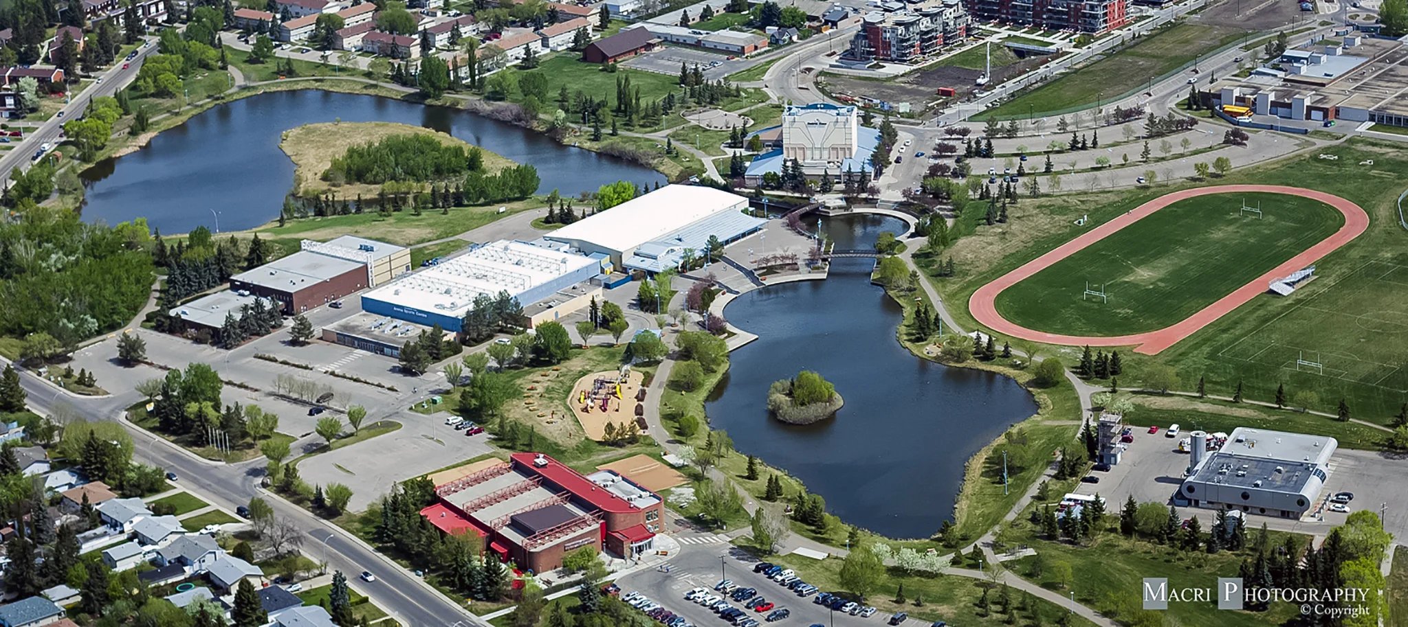 Aerial view of a park with two ponds, playground, buildings, running track, and parking lots.