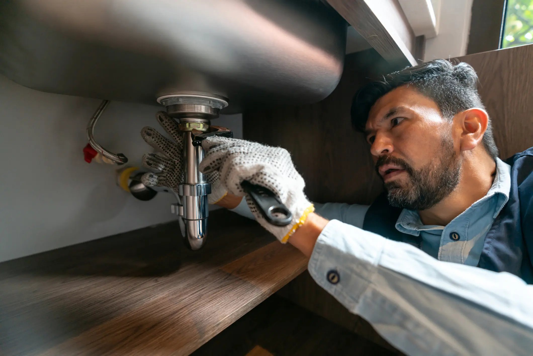 Man wearing gloves fixing plumbing under a kitchen sink with a wrench in a wooden cabinet.