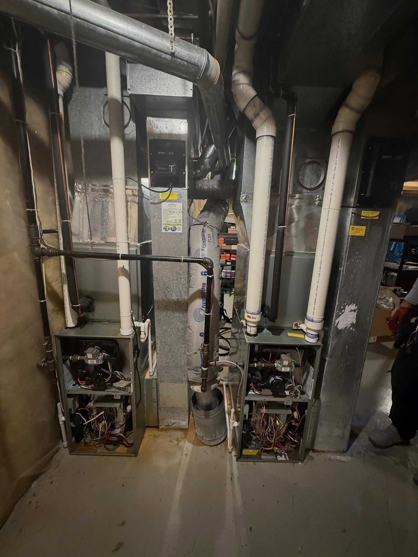 Two open HVAC units with exposed wiring, connected to ventilation pipes in a basement.