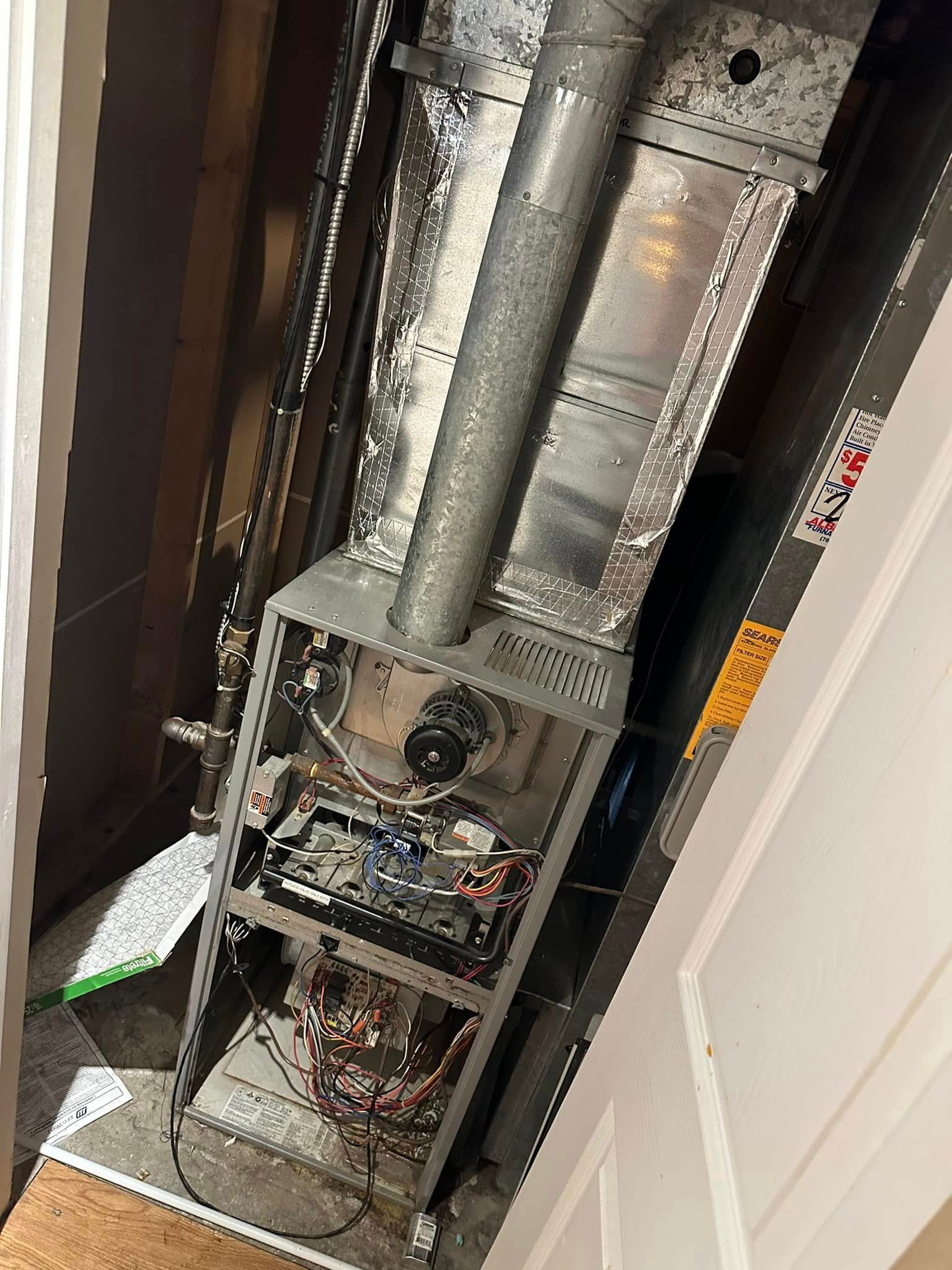 Open furnace unit with visible wiring and pipes inside a small utility closet.