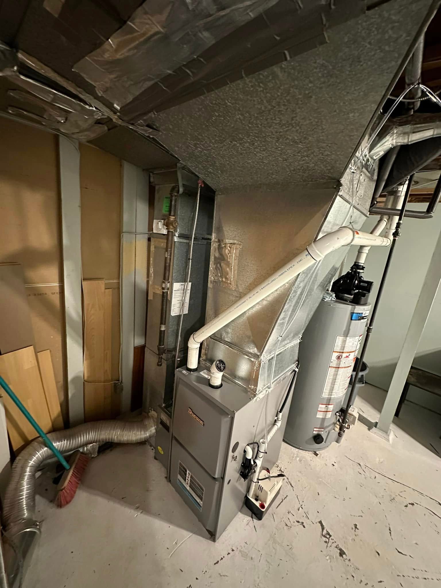 Basement furnace and water heater unit with ducts, pipes, and a broom on concrete floor.