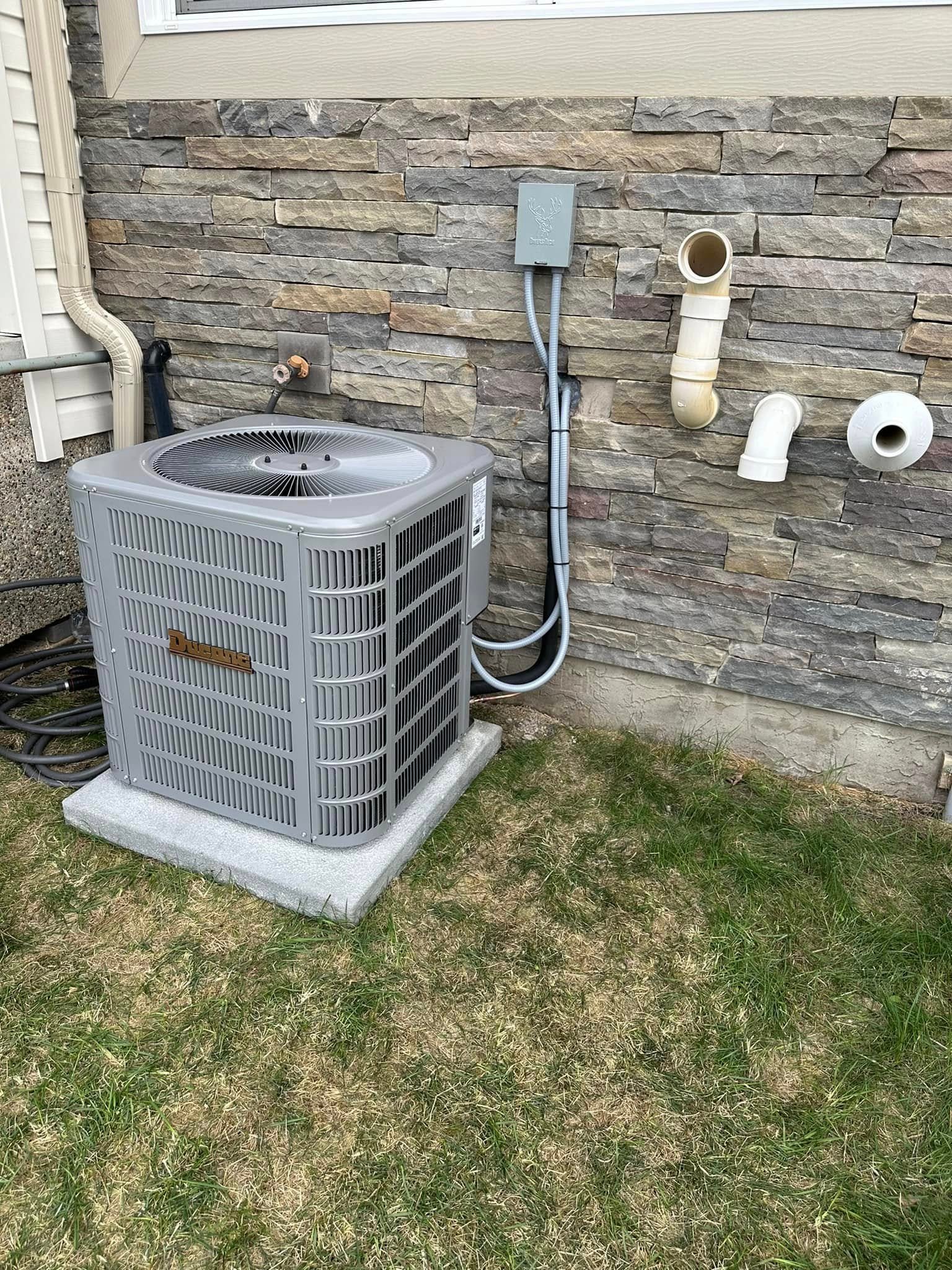 Gray outdoor air conditioning unit next to a stone wall with pipes and wiring attached.