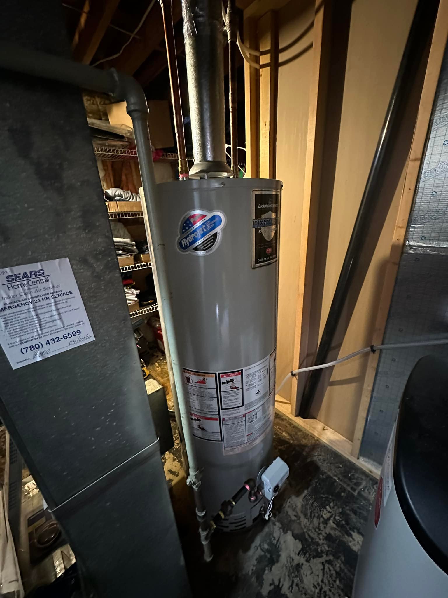 Tall cylindrical water heater with pipes and warning labels in a dimly lit basement.
