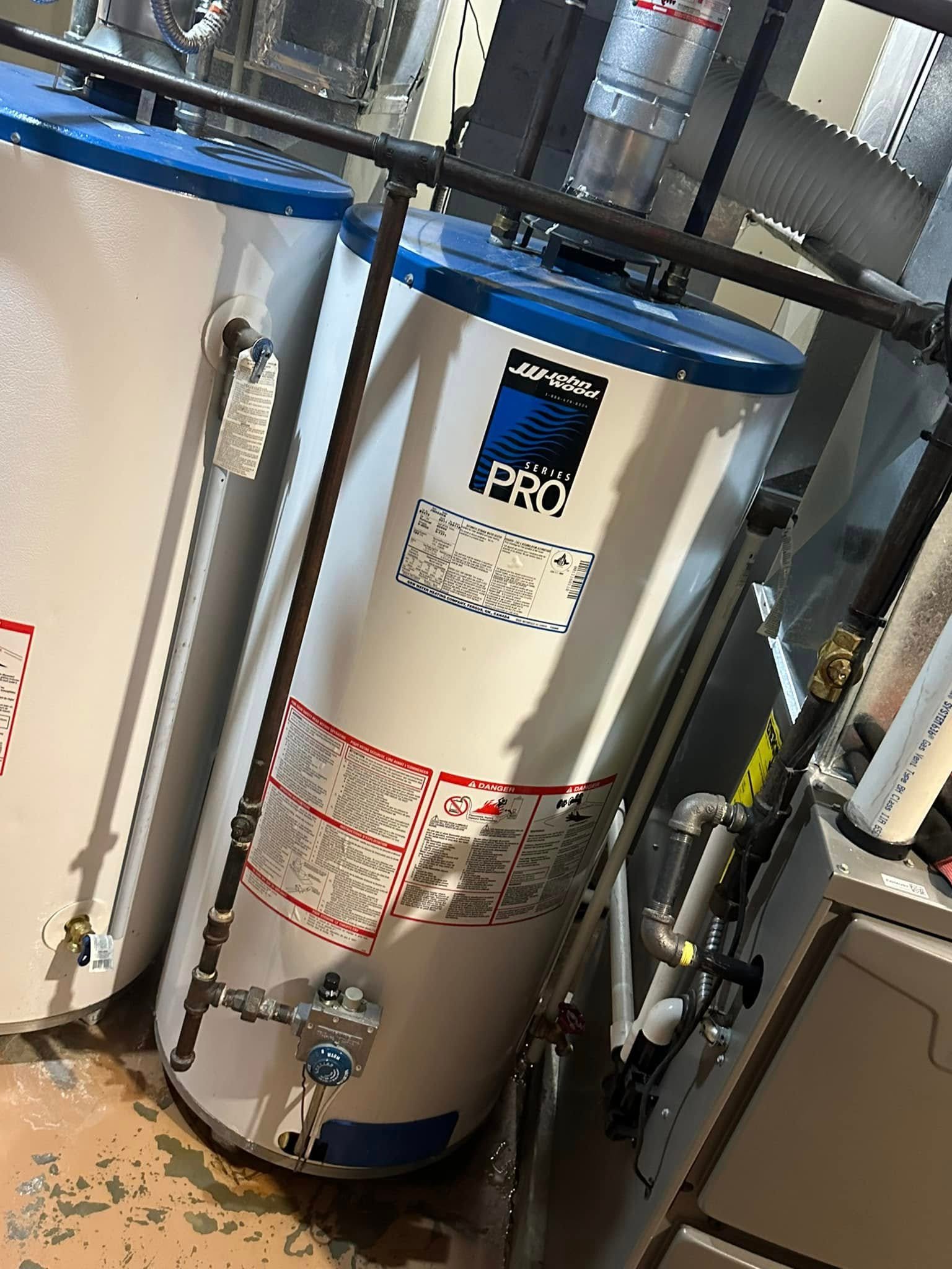 Two John Wood Pro Series water heaters with blue tops, pipes, and warning labels in a utility room.