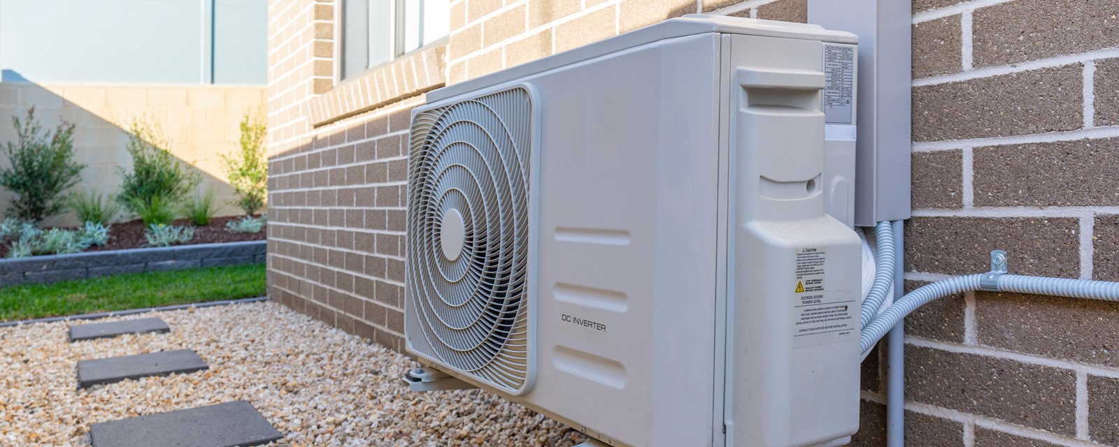 Outdoor DC inverter air conditioning unit mounted on exterior brick wall with gravel ground below.