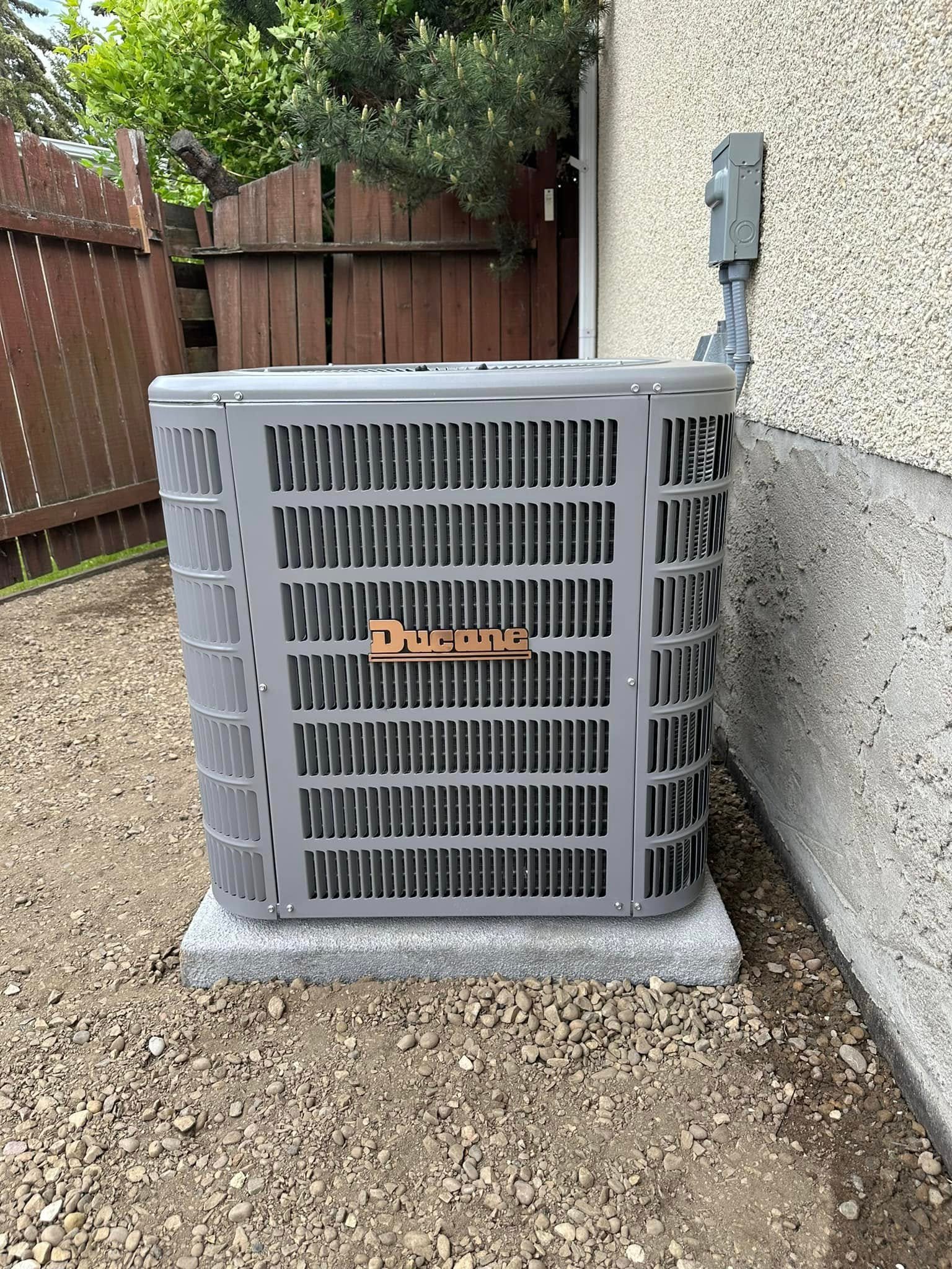 Gray Ducane air conditioning unit installed on a concrete slab next to a house wall and wooden fence.