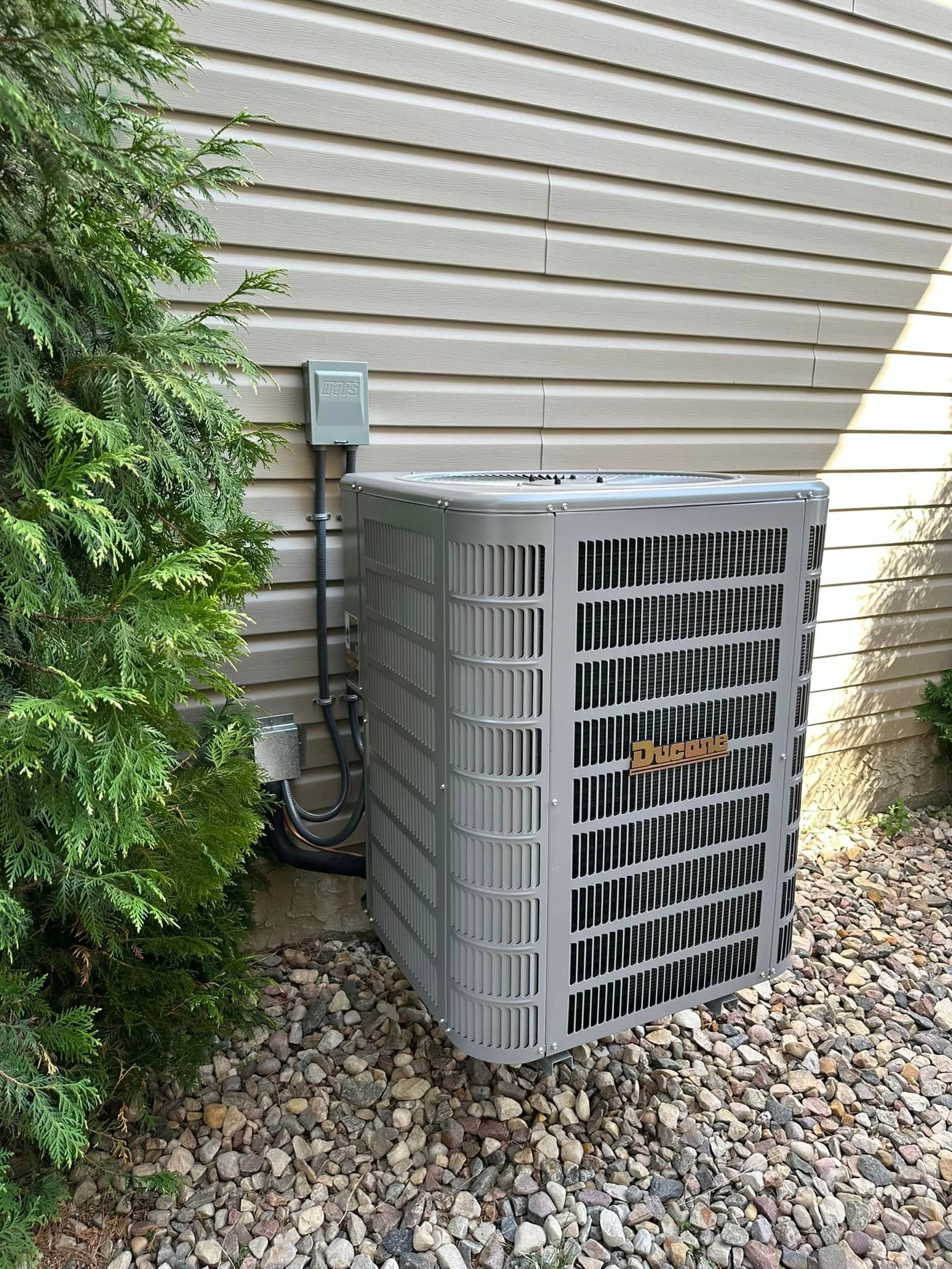 Ducane air conditioning unit installed on rock-covered ground beside a beige house with siding.