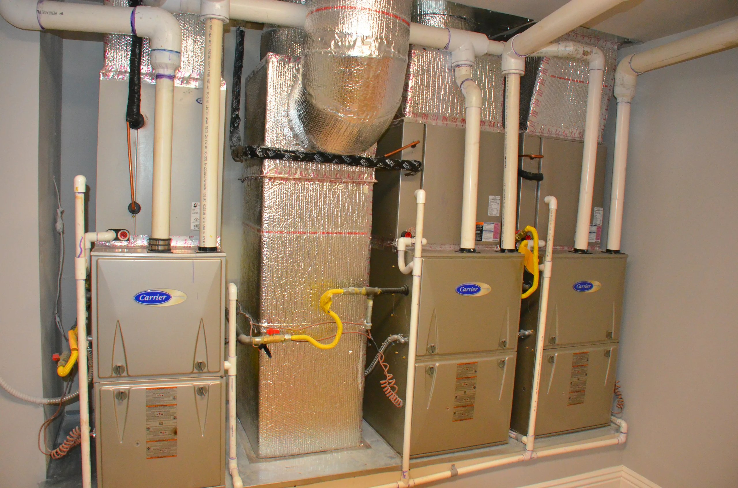 Three beige Carrier HVAC units with white pipes and insulated ductwork in a utility room.