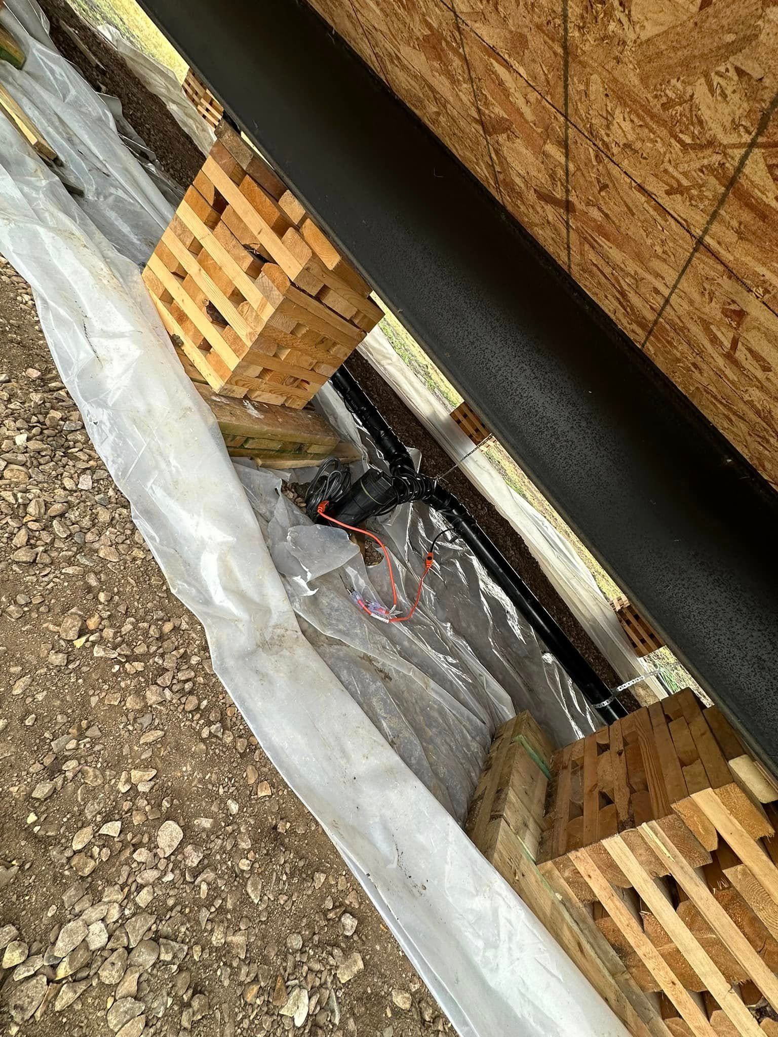 Black wrapped beam supported by wooden blocks over plastic sheeting and gravel on the ground.