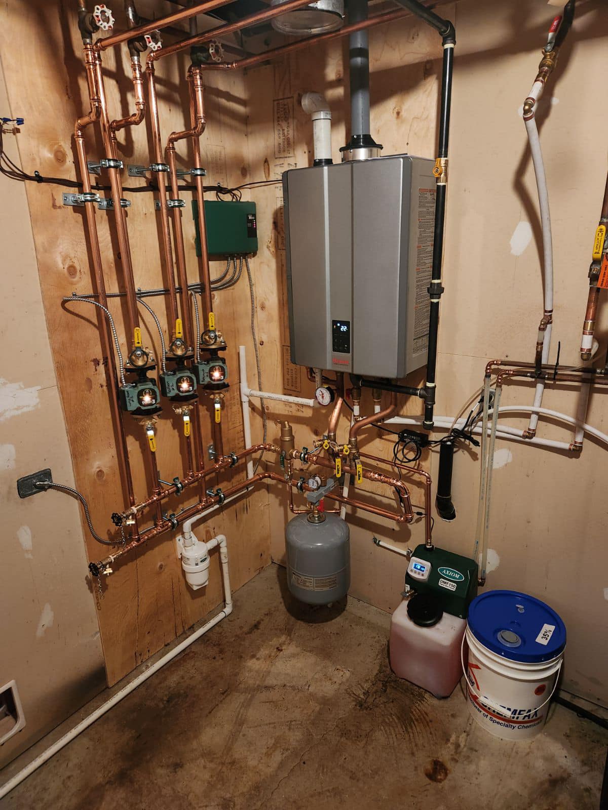 Indoor utility room with a boiler system, copper pipes, pressure tank, and chemical containers on concrete floor.