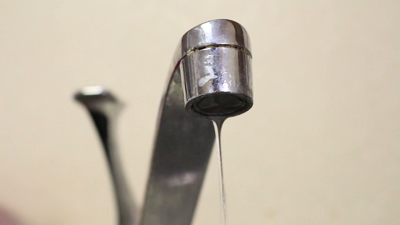 Metal faucet with a slow stream of water flowing against a plain beige background.