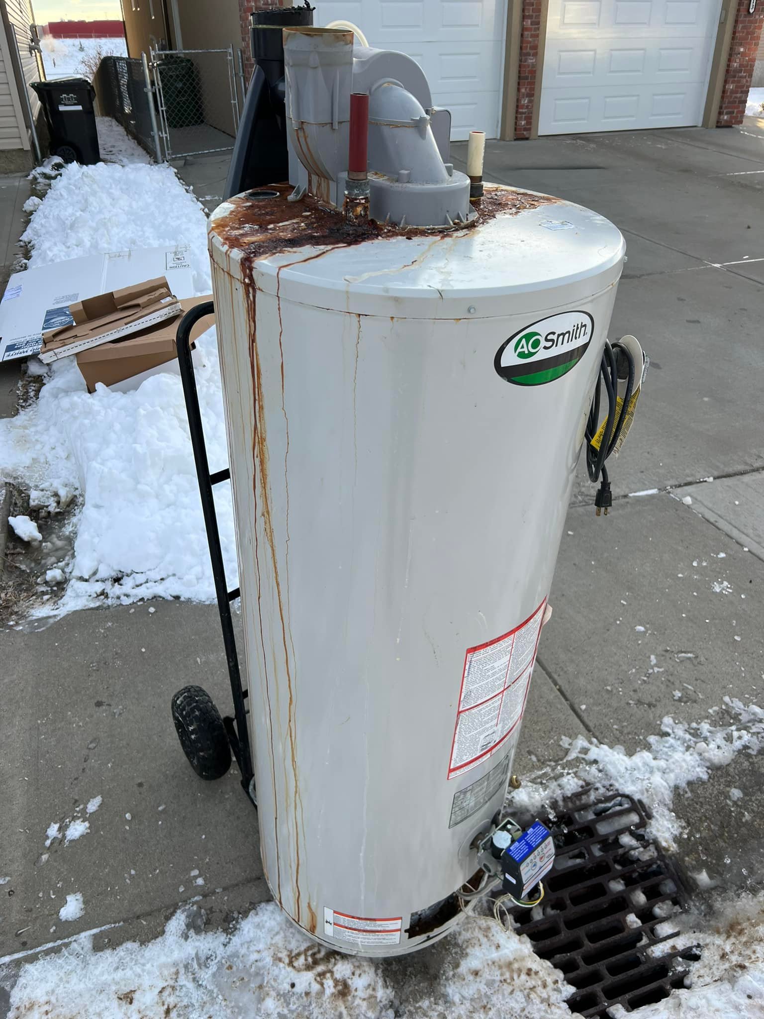 Rusty white AO Smith water heater on a dolly outside on snowy pavement near a drain.
