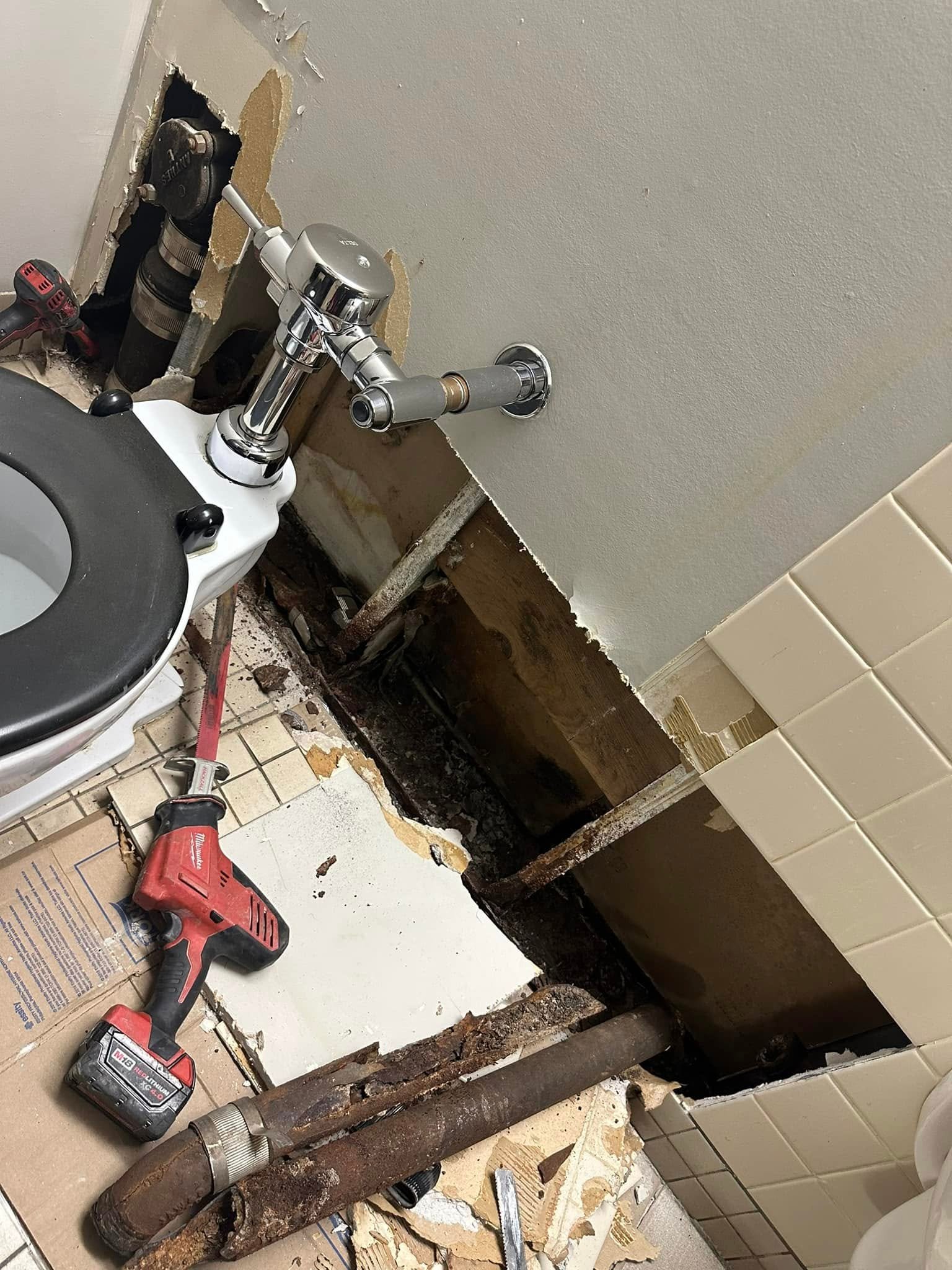 Bathroom under repair showing exposed rusty pipes, removed wall panel, toilet, and a red cordless drill on tiled floor.