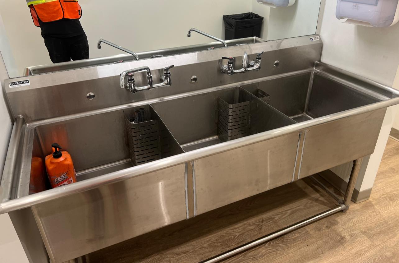 Three-compartment stainless steel handwashing sink with soap dispenser and metal utensil holders.