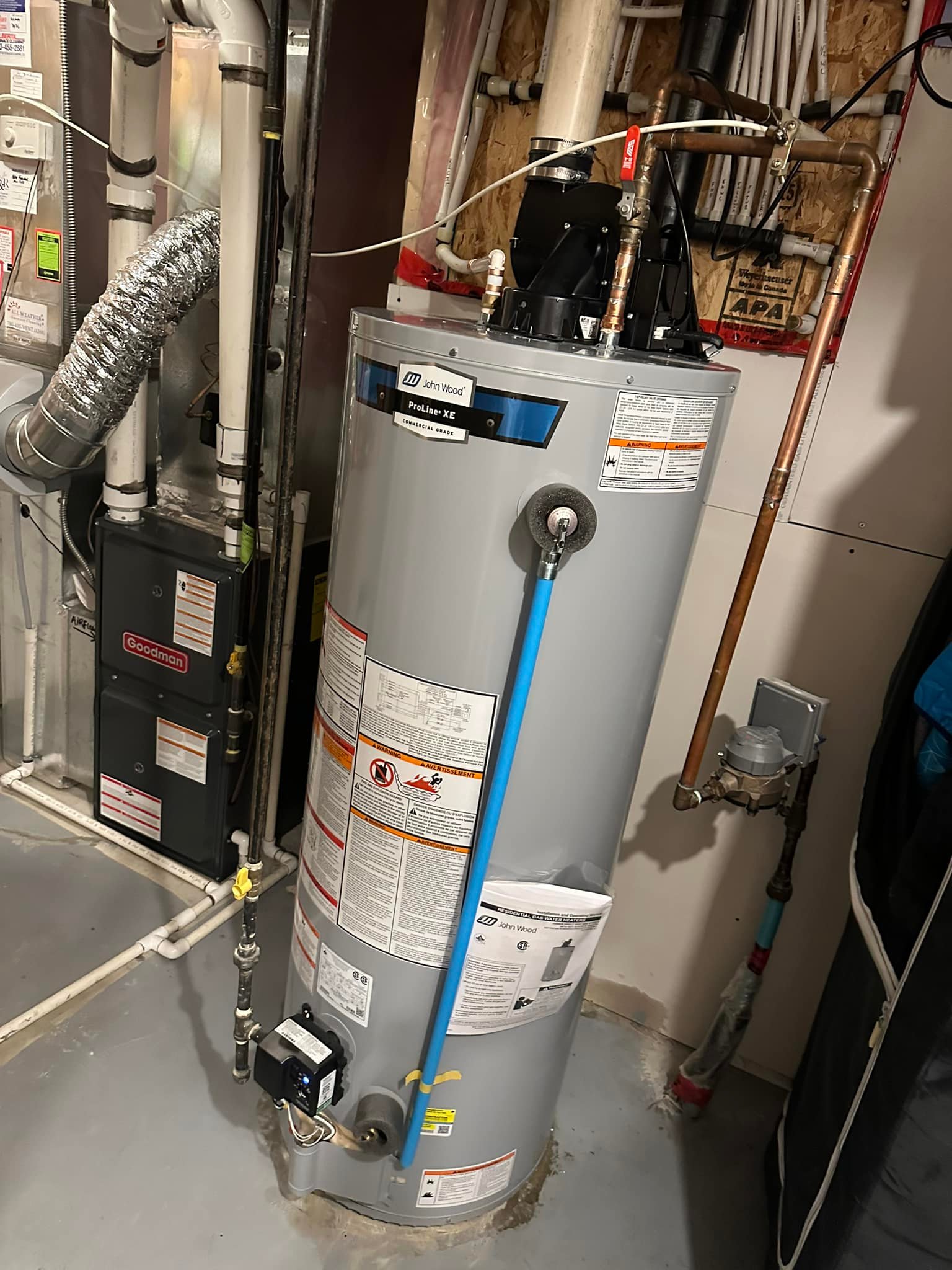 John Wood ProLine XE gray water heater with pipes and warning labels in a basement utility area.