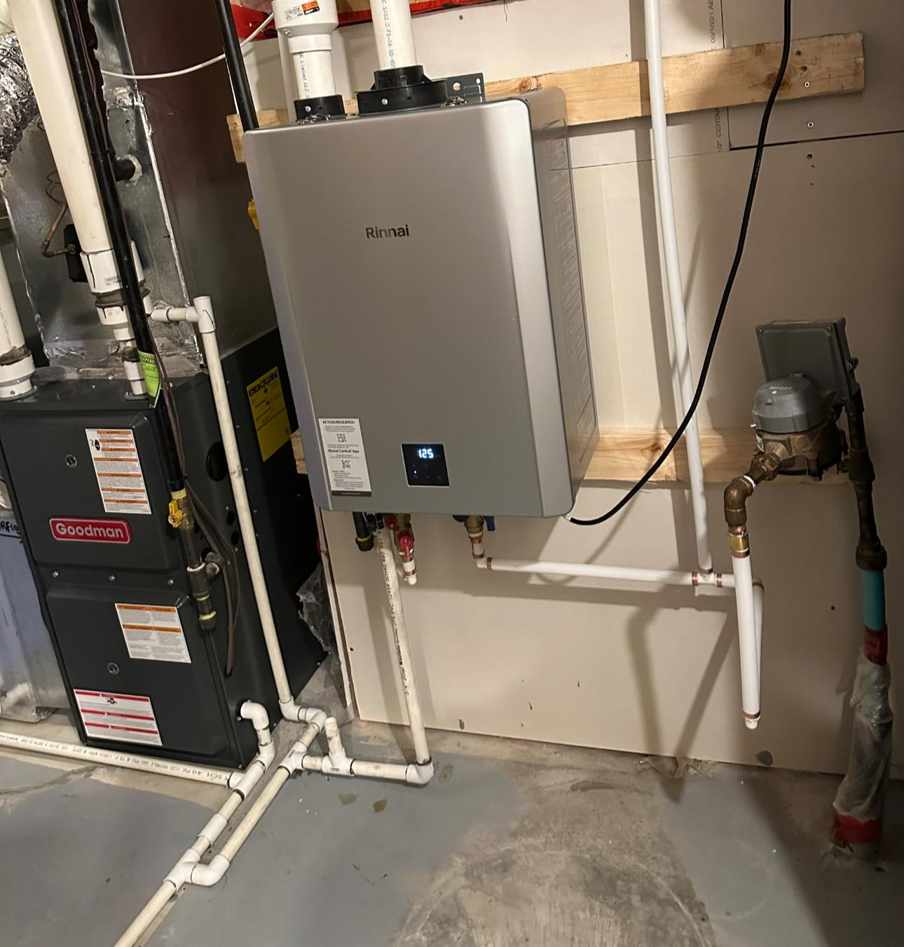 Basement utility area with a wall-mounted Rinnai tankless water heater and a Goodman furnace connected with white pipes.