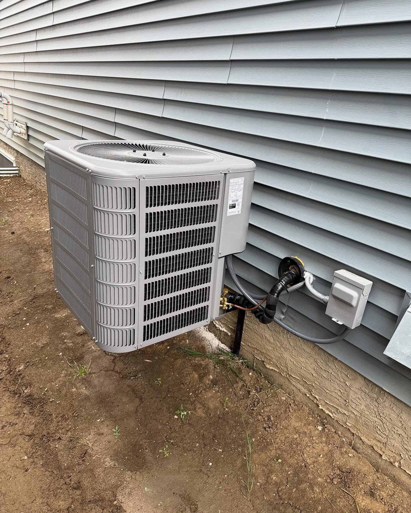Modern gray outdoor HVAC unit installed next to a light blue vinyl-sided house with exposed dirt ground.