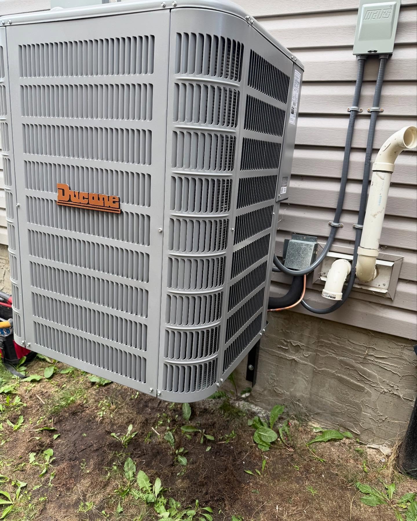 Gray Ducane air conditioning unit mounted outside a house with siding and pipes attached.