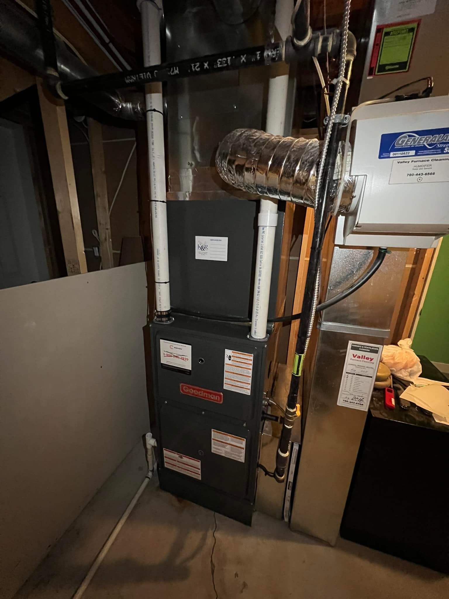 Goodman HVAC furnace unit in a basement with pipes, ductwork, and utility labels visible.