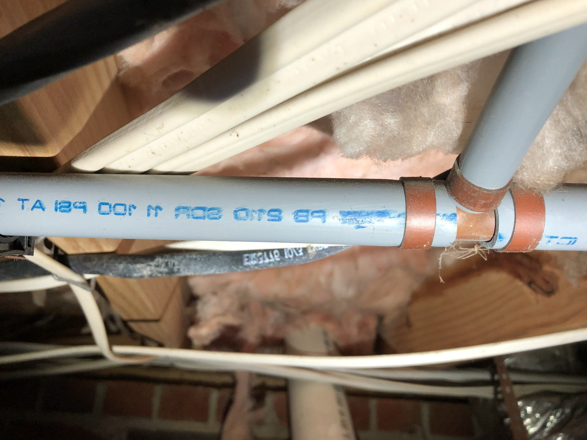 Gray plumbing pipes joined with copper clamps among wood and insulation in a basement ceiling.