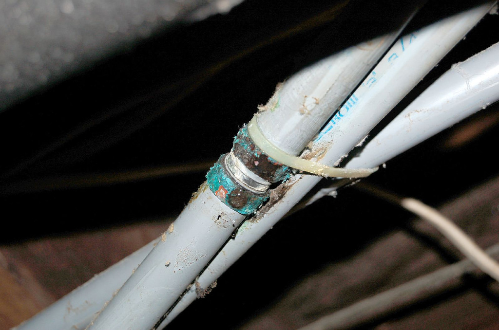 Close-up of corroded metal pipe fitting connecting white PVC pipes with a plastic zip tie.