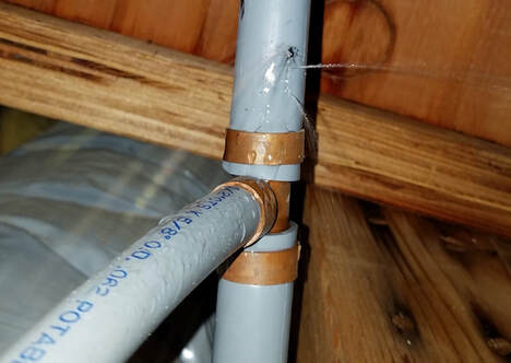 Gray PVC plumbing pipes connected with copper fittings inside wooden ceiling framing.