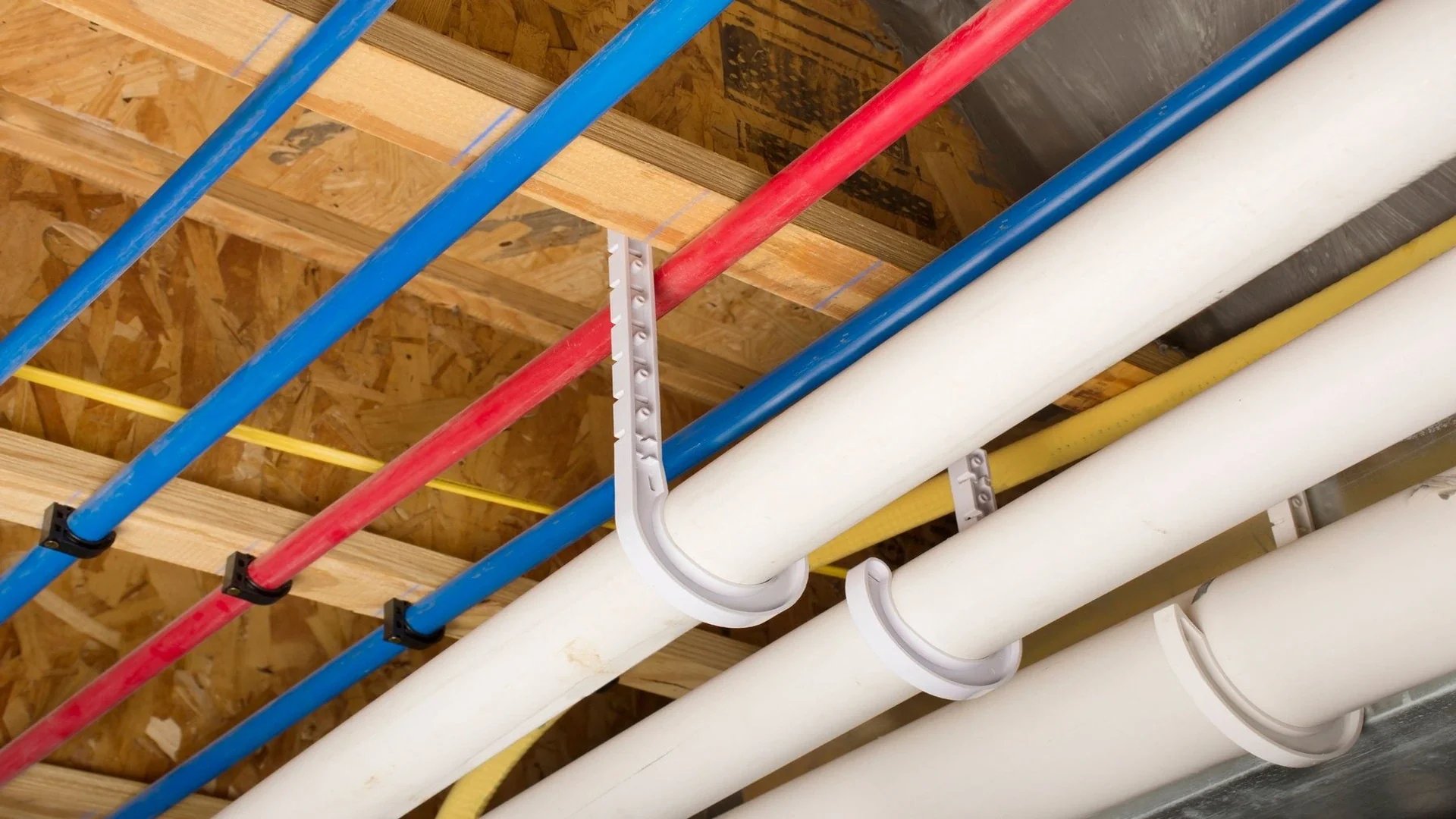 Red, blue, yellow, and white pipes secured with pipe clamps on a wooden ceiling structure.
