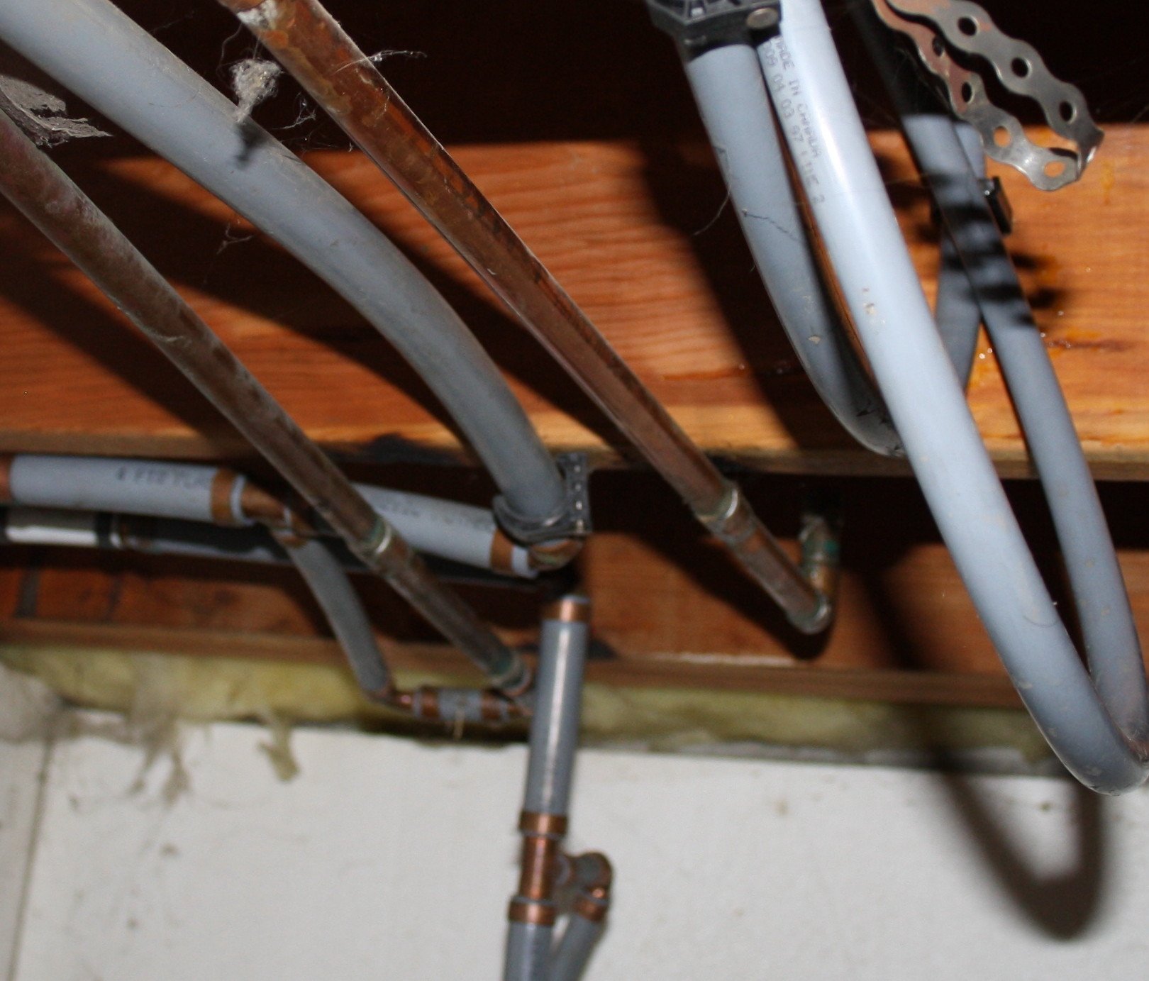 Copper and plastic water pipes running beneath wooden beams with some insulation above a concrete wall.