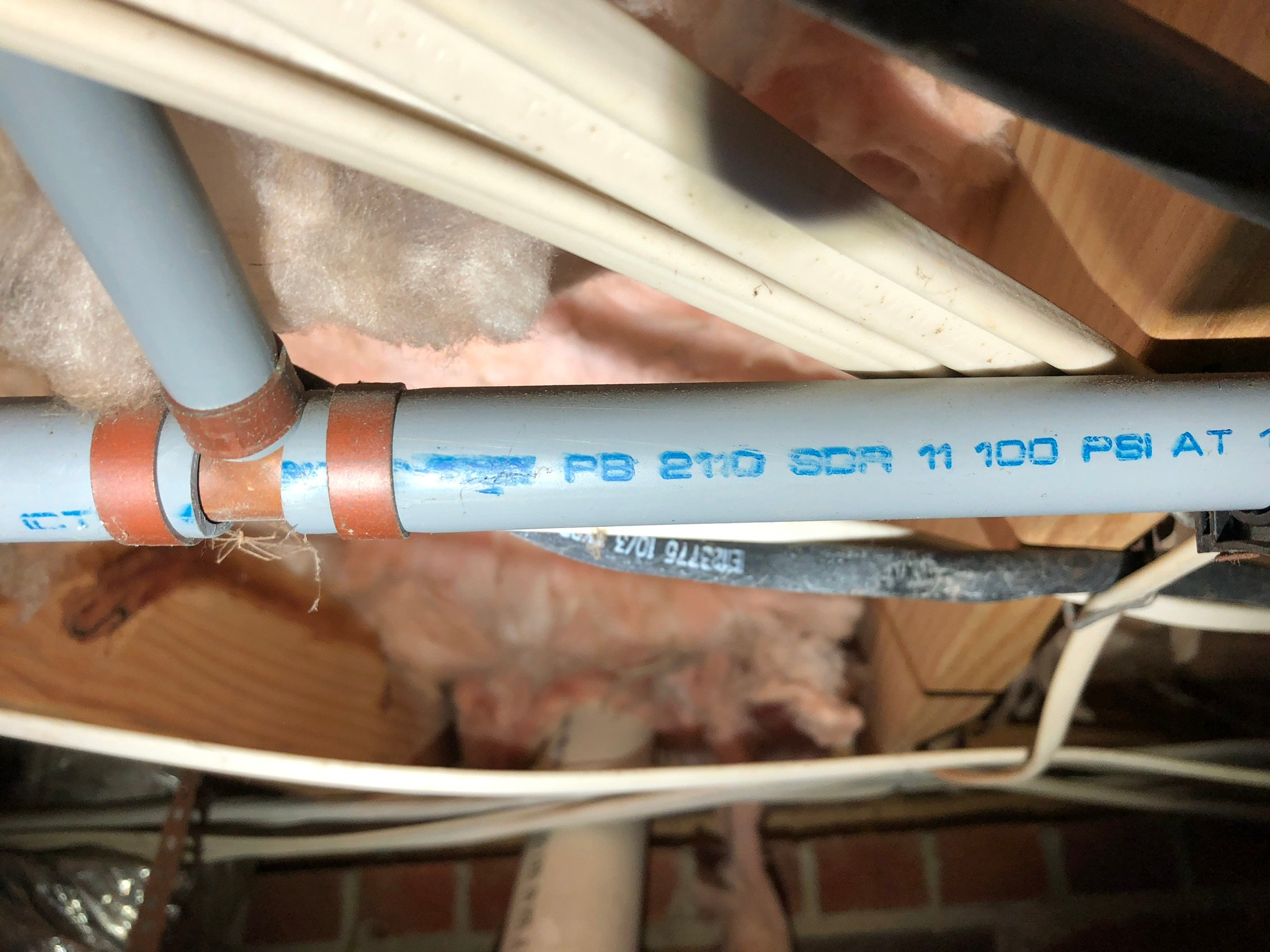 Close-up of gray plumbing pipes with red clamps and pink insulation in a wooden crawl space.