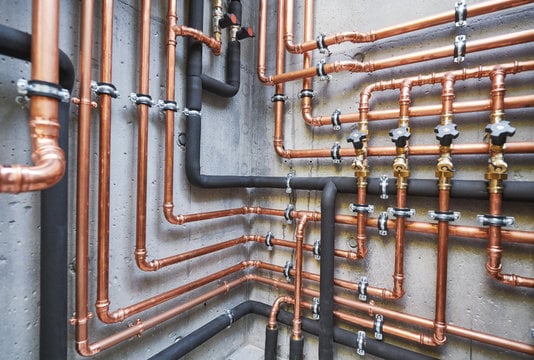 Copper and black pipes arranged along a concrete wall with valves and clamps in an organized plumbing system.