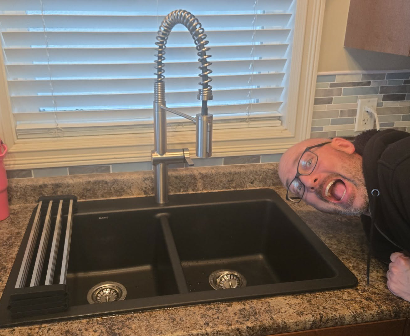 Man with glasses smiling widely next to a modern black kitchen sink with a tall coiled faucet.